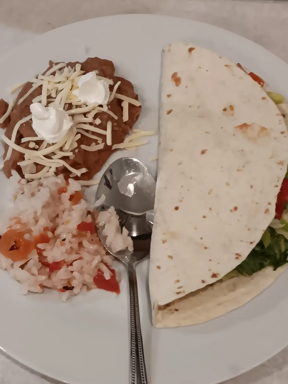 A plate of food featuring a folded burrito on the right, refried beans topped with shredded cheese and sour cream on the…