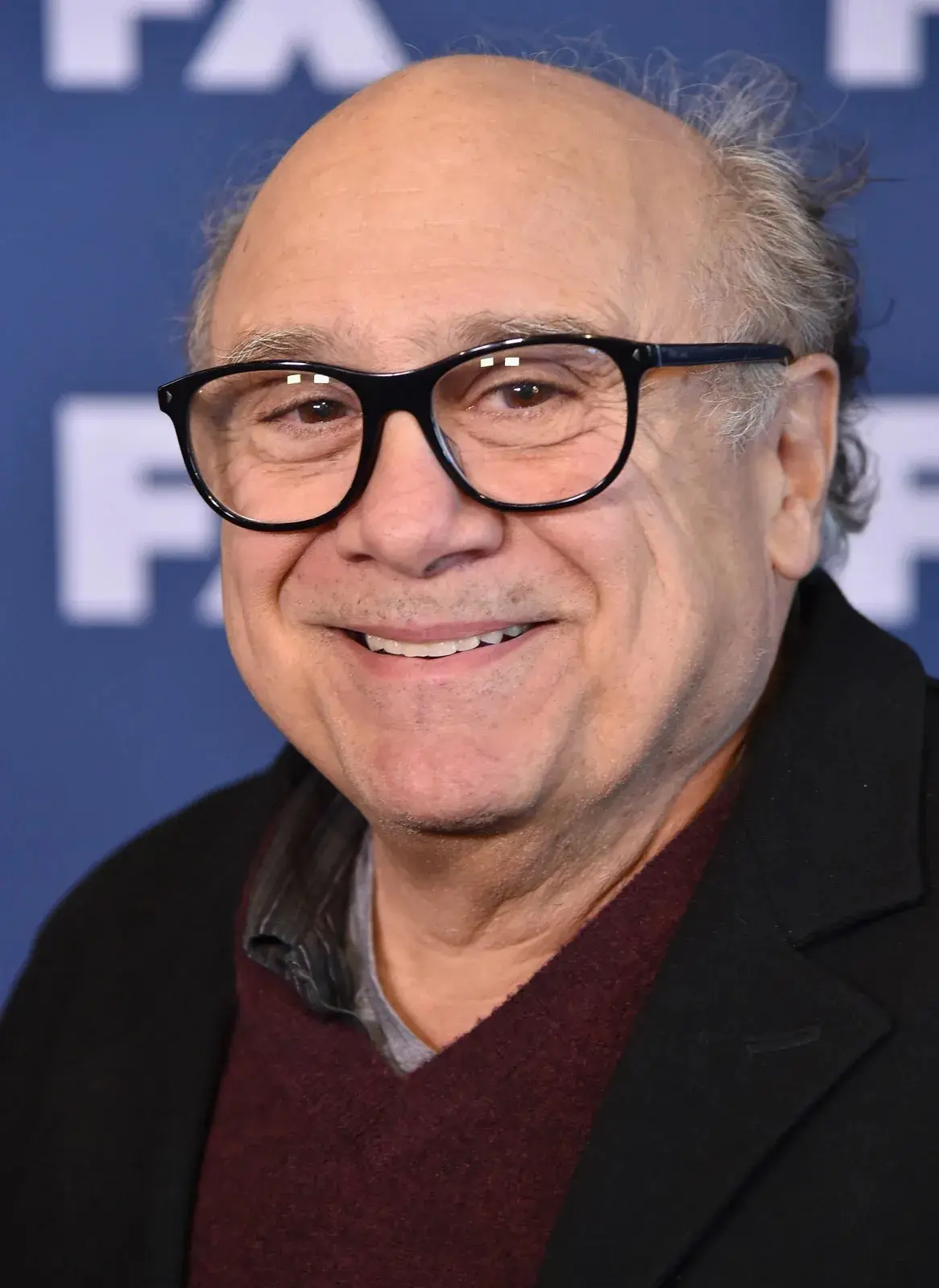 A close-up photo of actor Danny DeVito smiling while wearing large black-rimmed glasses, a dark coat, and a maroon sweater…