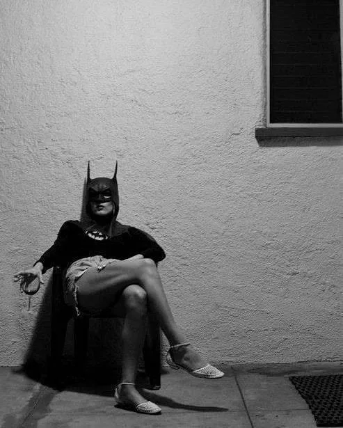 A black-and-white photo shows a woman in a Batman costume sitting on a chair against a textured wall, wearing a short skirt…