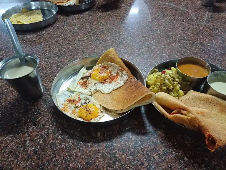 A metal plate holds a dosa topped with two sunny-side-up eggs seasoned with red chili powder and black pepper, alongside a…