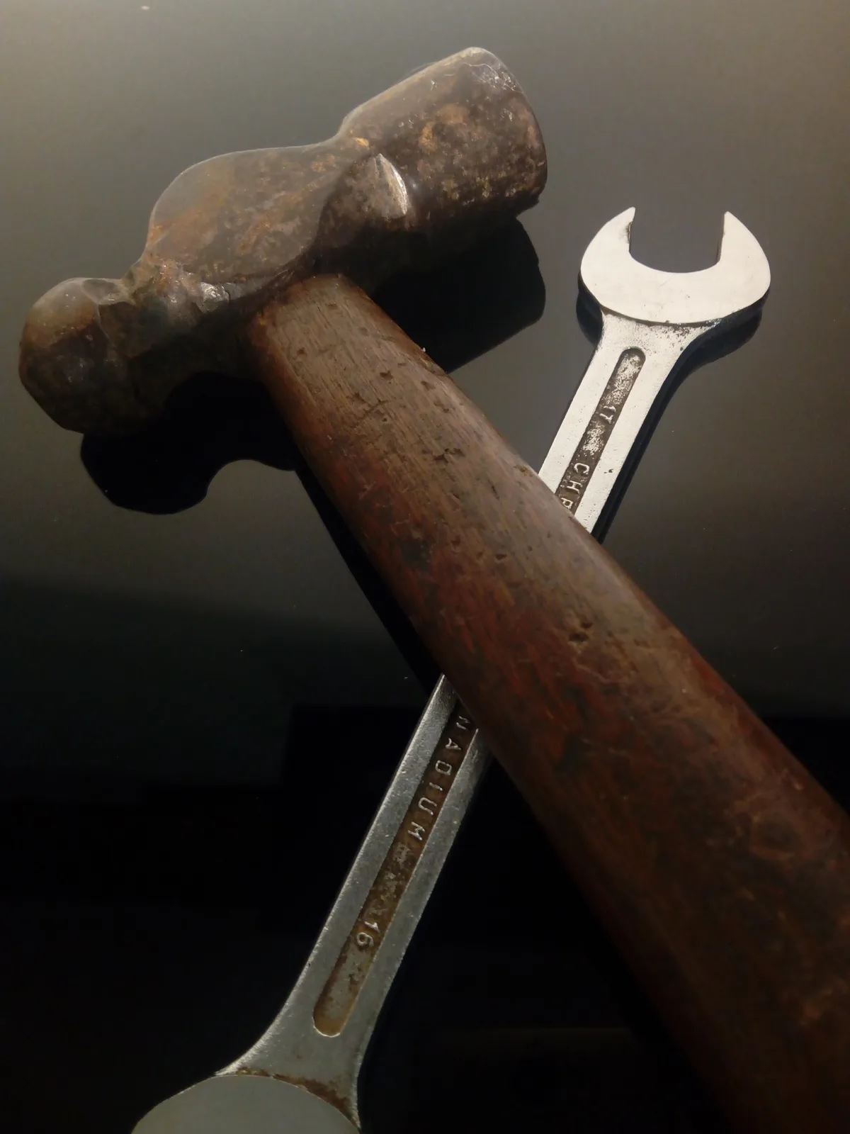 A close-up photo shows a well-worn claw hammer with a wooden handle and a metal wrench lying beside it on a dark reflective…