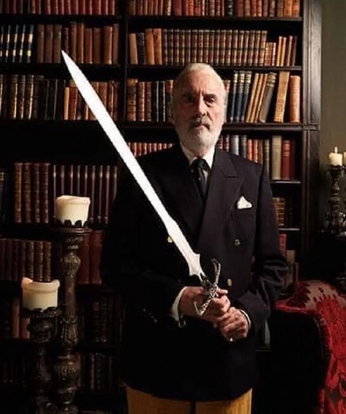 An elderly man with a white beard, wearing a dark formal suit and tie, stands in a library filled with bookshelves. He holds…