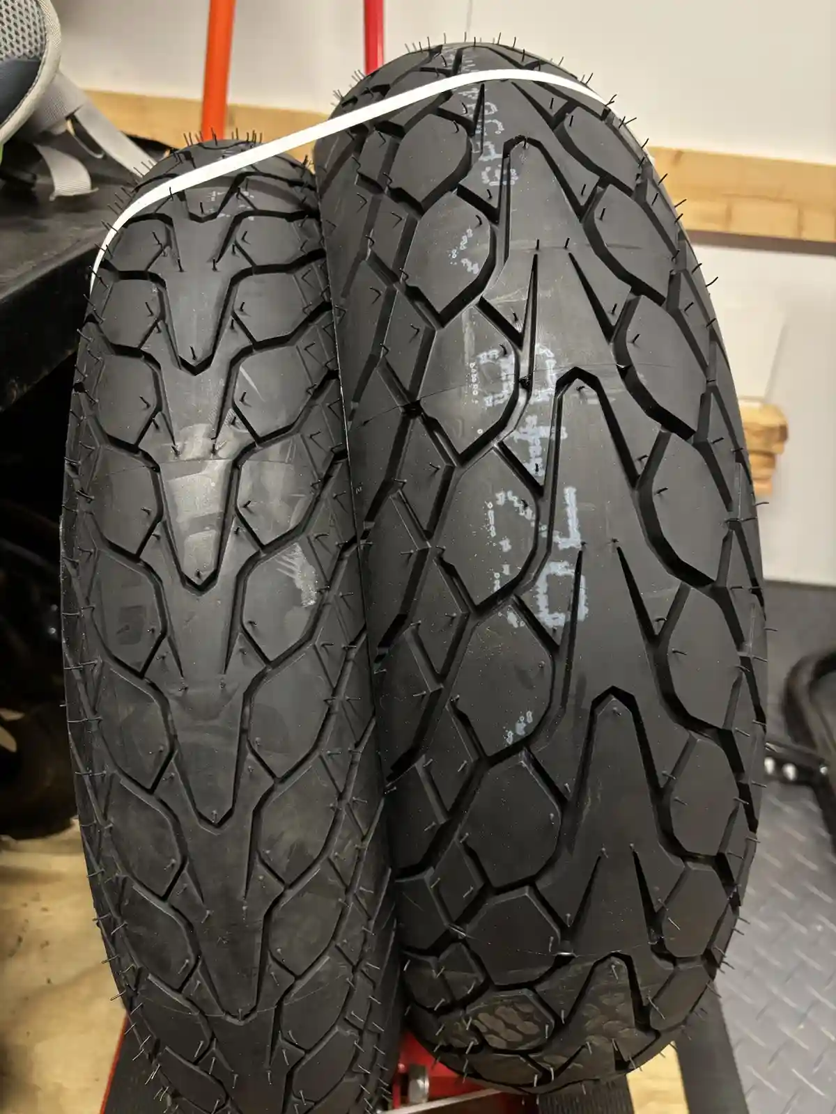 Two black motorcycle tires with aggressive tread patterns are shown side by side, both embedded with numerous small nails or…
