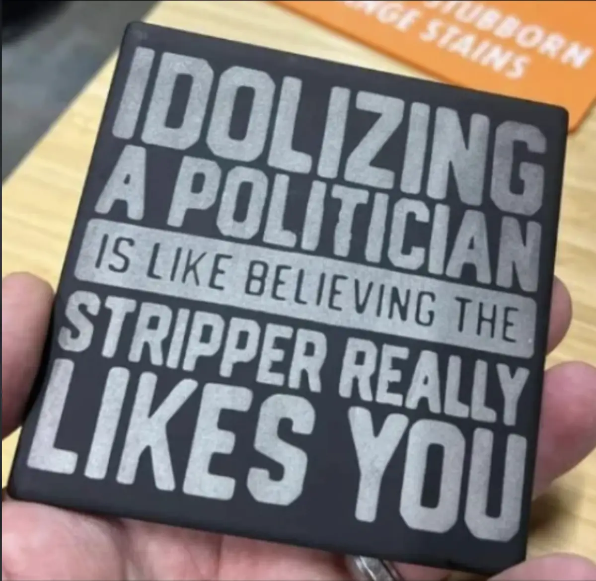 A hand holds a black square coaster with bold white text that reads 'IDOLIZING A POLITICIAN IS LIKE BELIEVING THE STRIPPER…