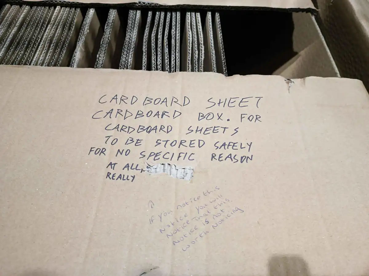 A close-up photo of a cardboard box with handwritten text on its side. The text humorously describes the box as containing…