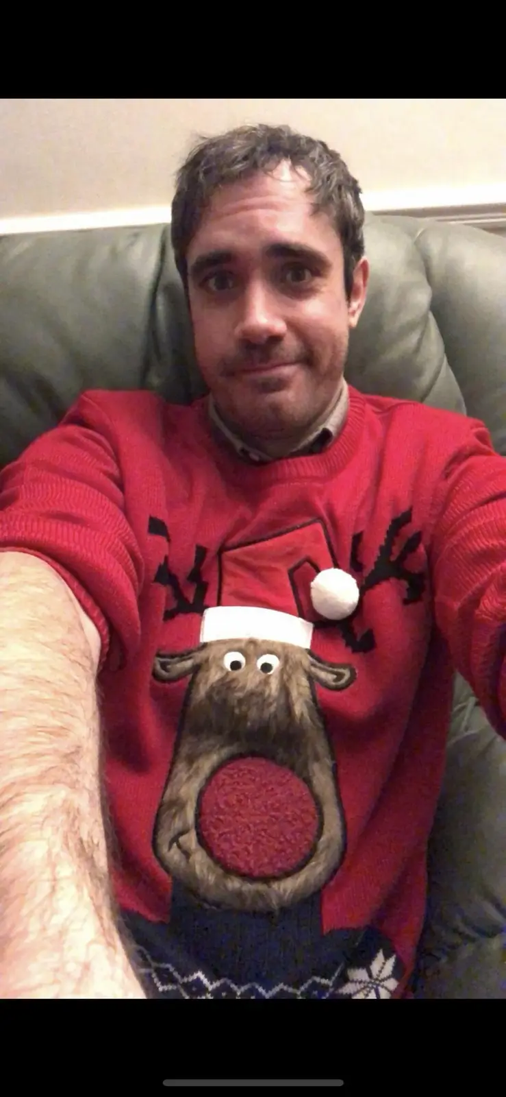 A man with short brown hair sits on a green couch, taking a selfie while wearing a red Christmas sweater featuring a large…
