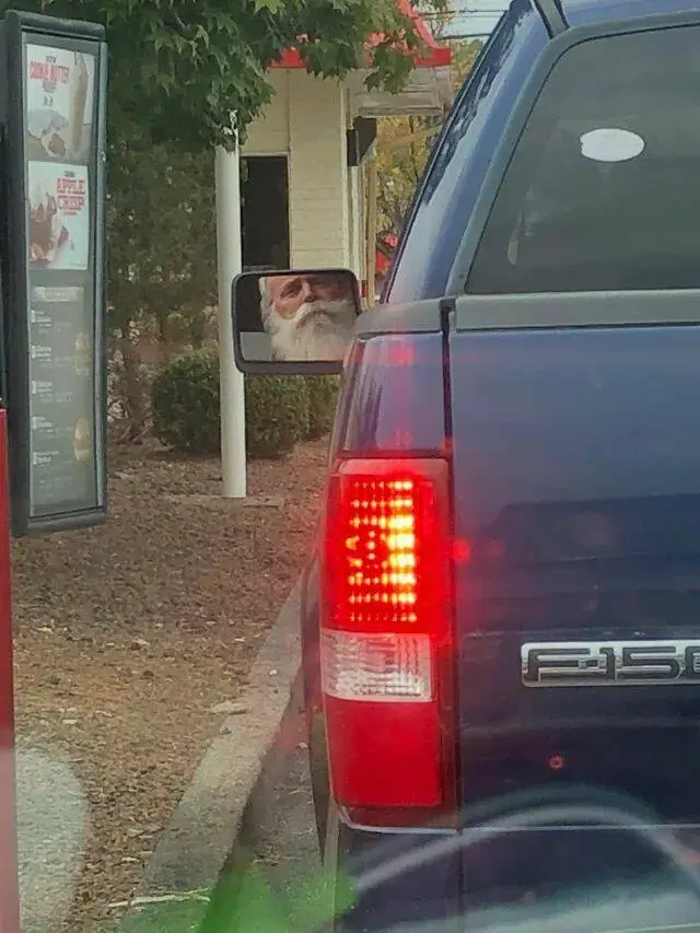 A dark blue Ford F-150 pickup truck is parked at a drive-thru, its rear passenger-side mirror reflecting the face of a man…