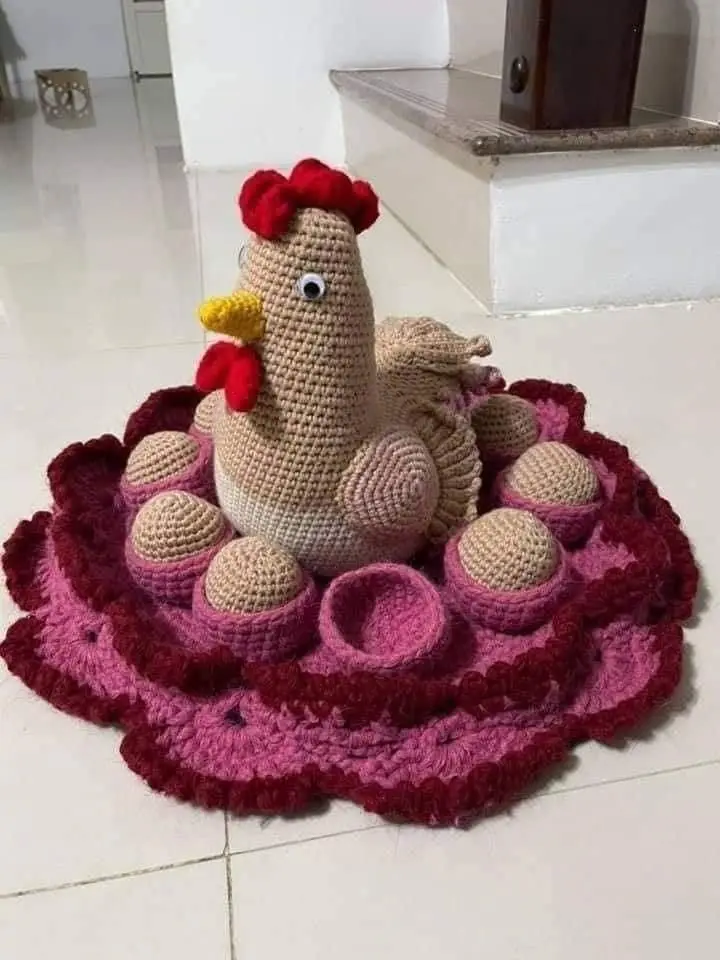 A handmade crochet chicken with a red comb and wattle sits on a pink and red crocheted nest, surrounded by several crochet…