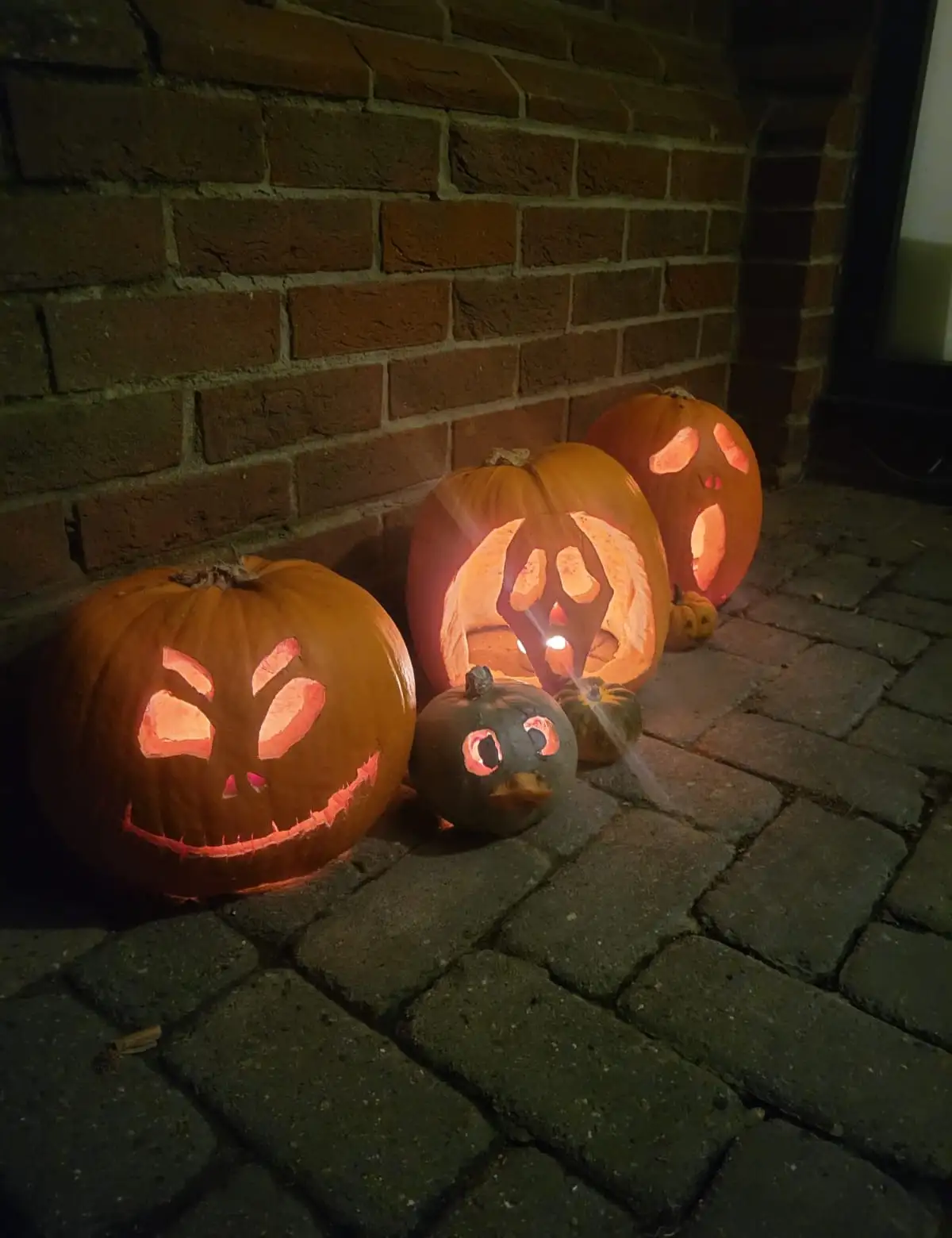 Several carved Halloween pumpkins with glowing faces sit on a stone patio against a brick wall. The smallest pumpkin, in the…
