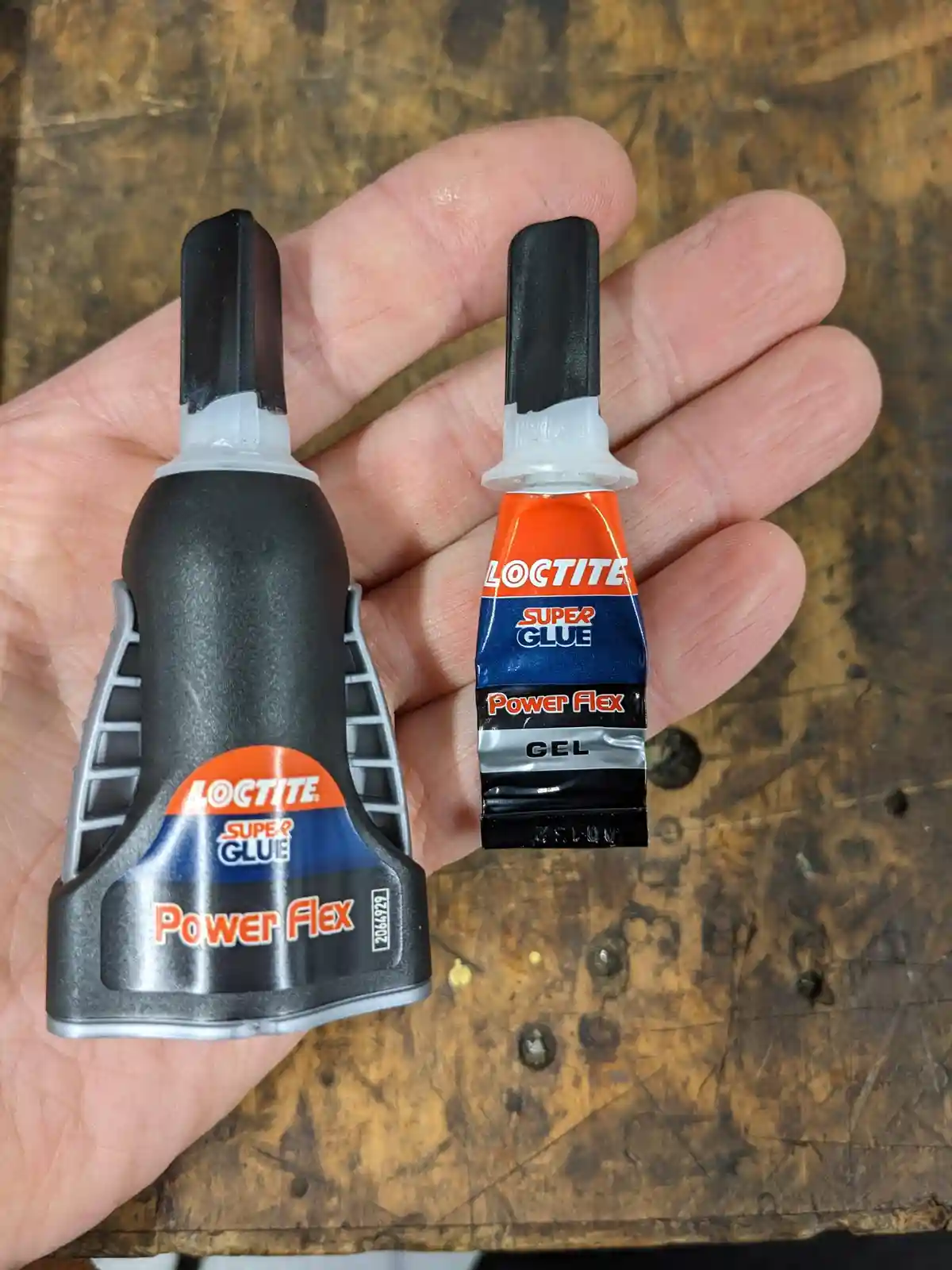 A hand holds two Loctite Super Glue Power Flex products side by side: a large, ergonomic black and silver glue gun on the…