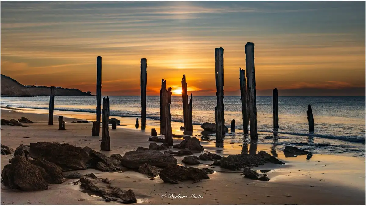 A serene beach sunset photo featuring weathered wooden pilings and rocks in the foreground, with the sun setting over the…