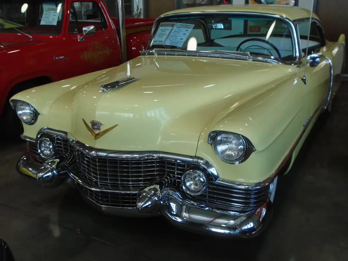 A pale yellow 1954 Cadillac Coupe DeVille is displayed in a showroom, with a price tag of $49,995 visible on the windshield…