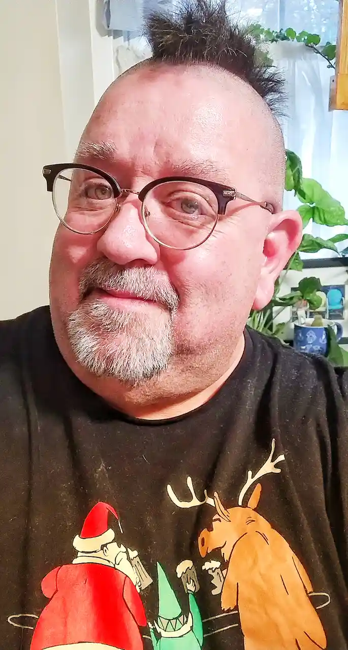 A man with glasses and a goatee wears a black t-shirt featuring cartoon illustrations of Santa Claus, a green goblin, and a…
