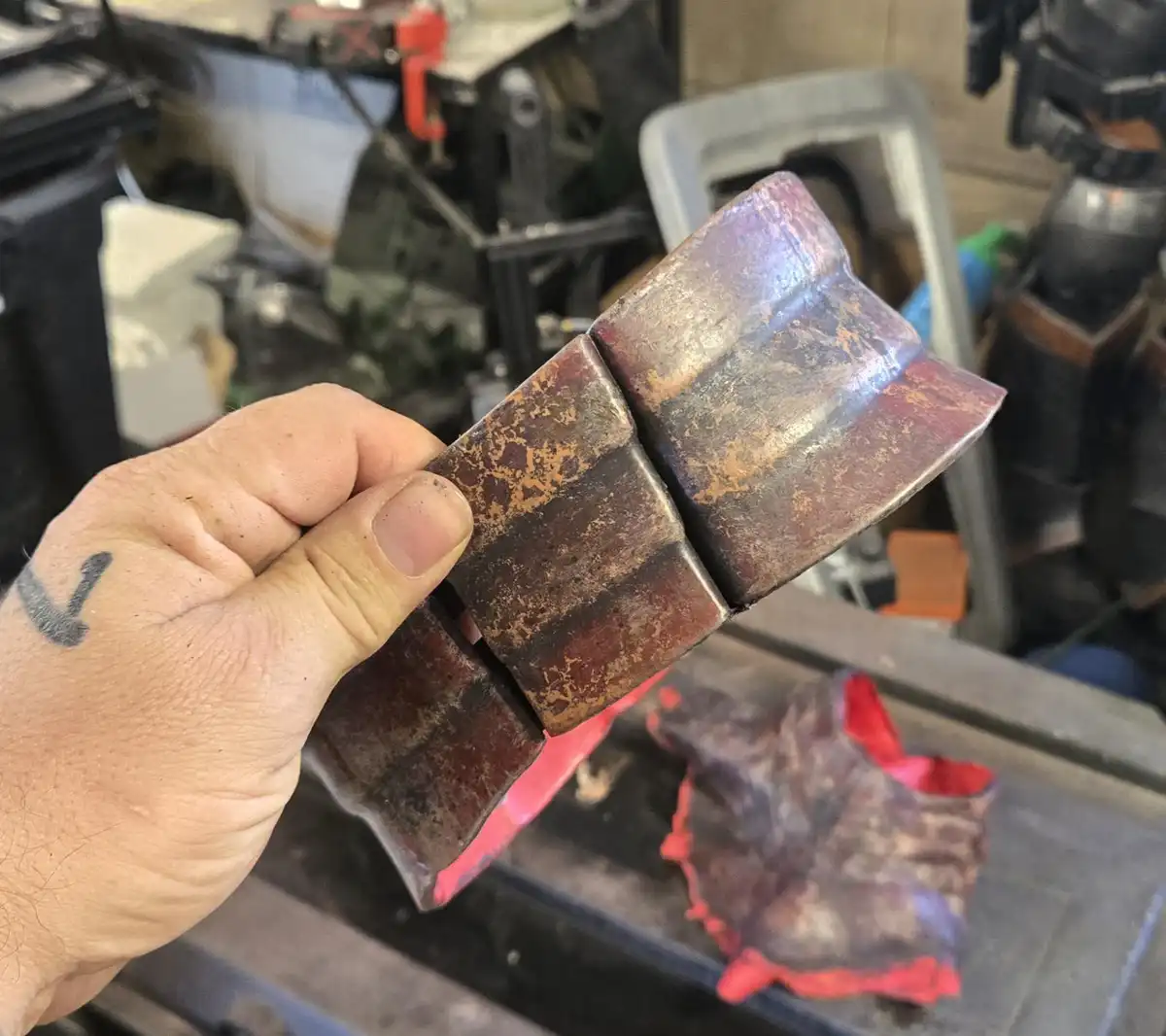 A hand holds a welded metal object with visible seams and rust, set against a cluttered workshop background. The overlaid…