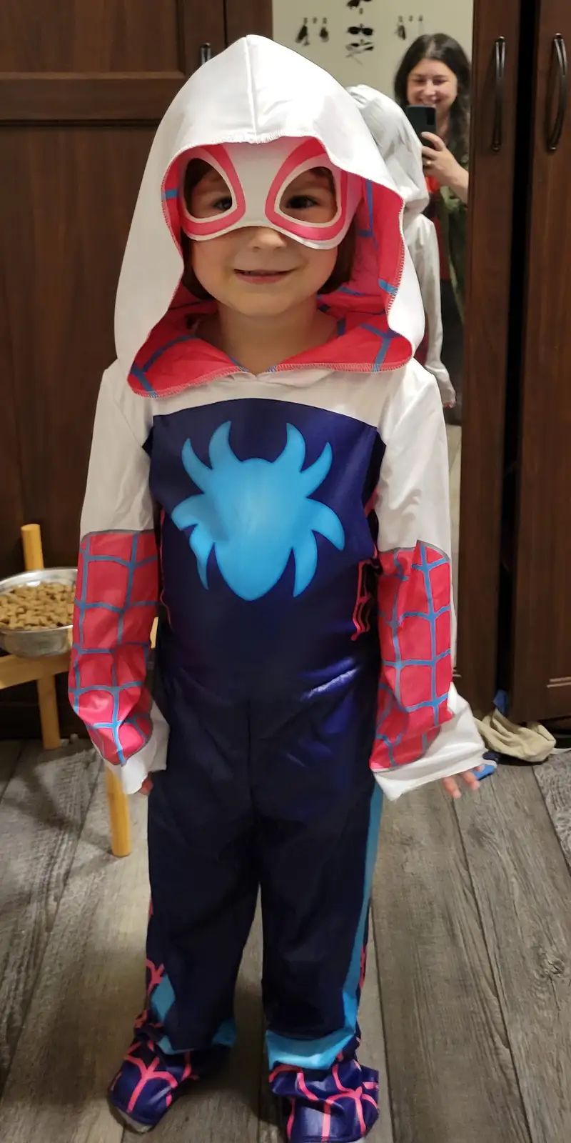 A young child wearing a full-body Spider-Gwen costume with a white hood, pink mask, blue spider emblem, and red…