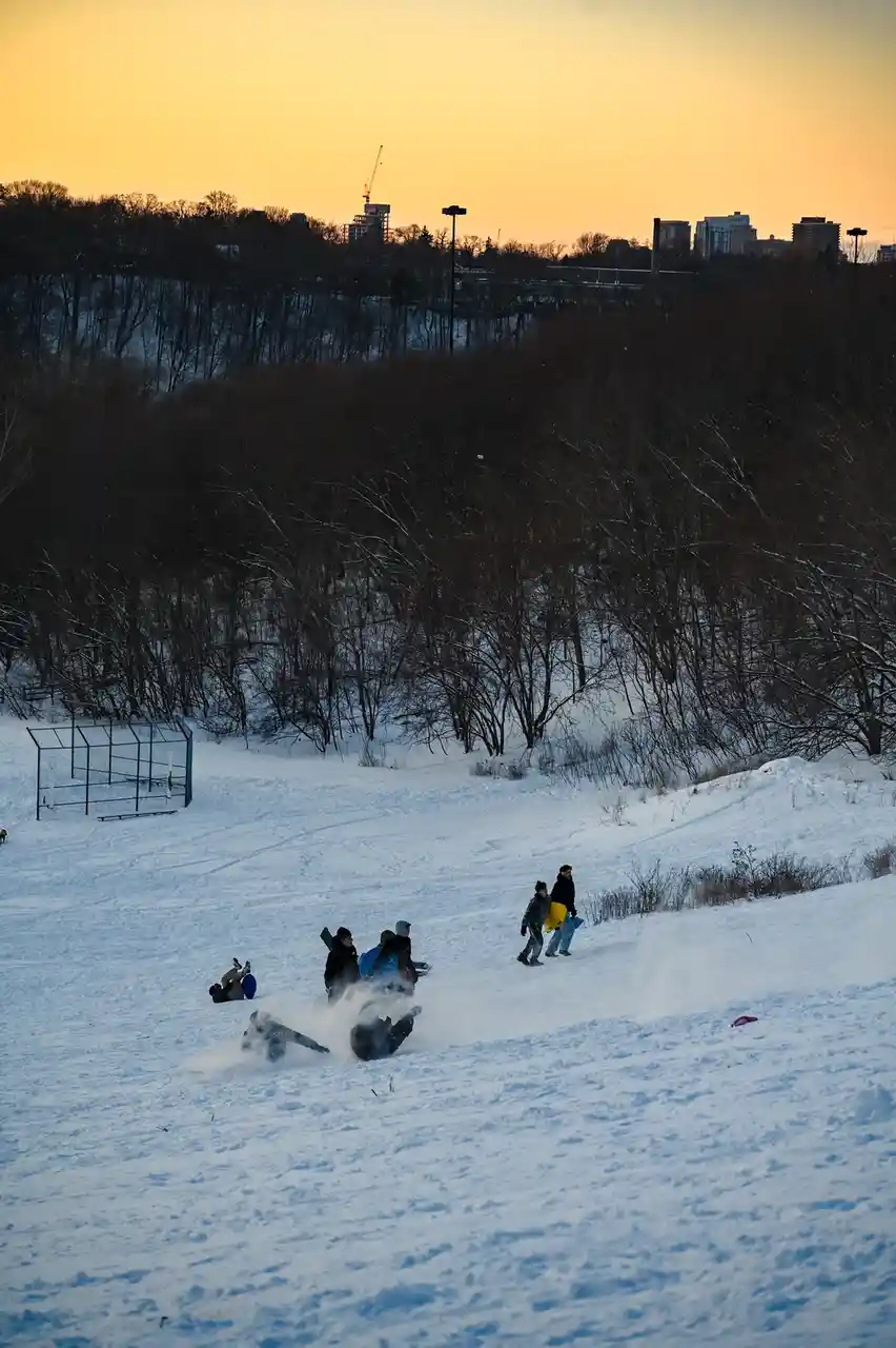 A snowy hill at sunset with people sledding and playing in the snow, some are sliding down while others stand nearby holding…