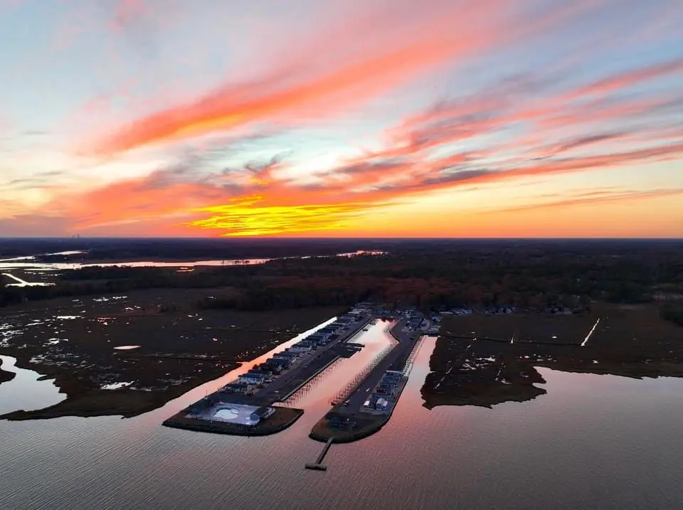 An aerial photo shows a serene harbor at sunset, with vibrant orange and pink clouds reflecting on the water. A long pier…