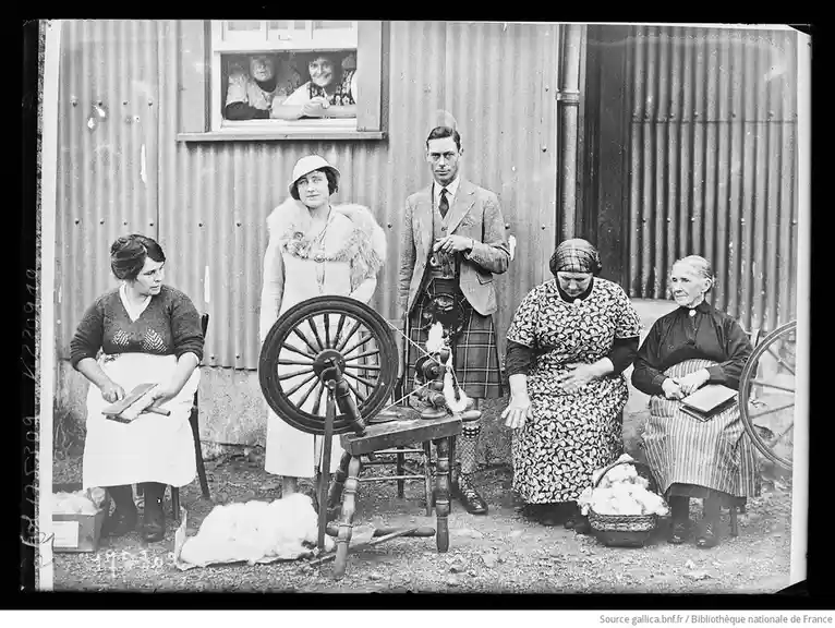 A vintage black-and-white photo shows a man in a kilt standing among several women with spinning wheels and wool. He holds a…