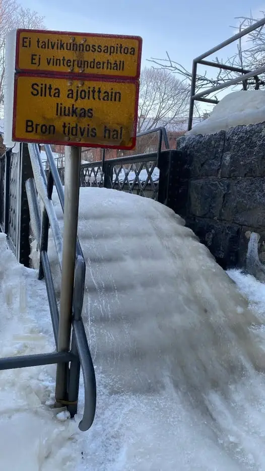 A photo of a snow-covered, icy bridge with a yellow warning sign in Finnish and Swedish. The sign warns that the bridge is…