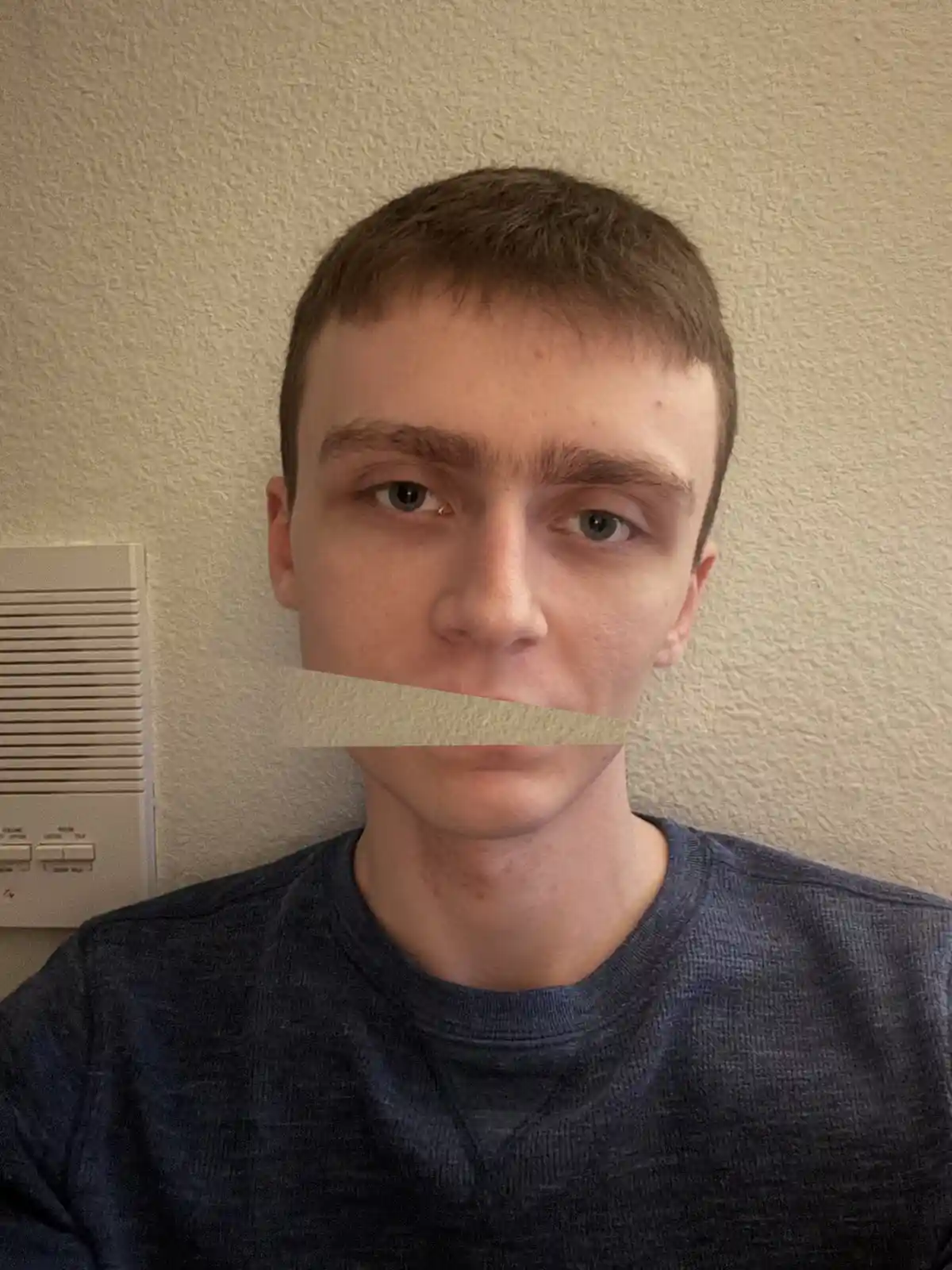A young man with short brown hair and blue eyes stares directly at the camera, his mouth covered by a beige tape overlay. He…