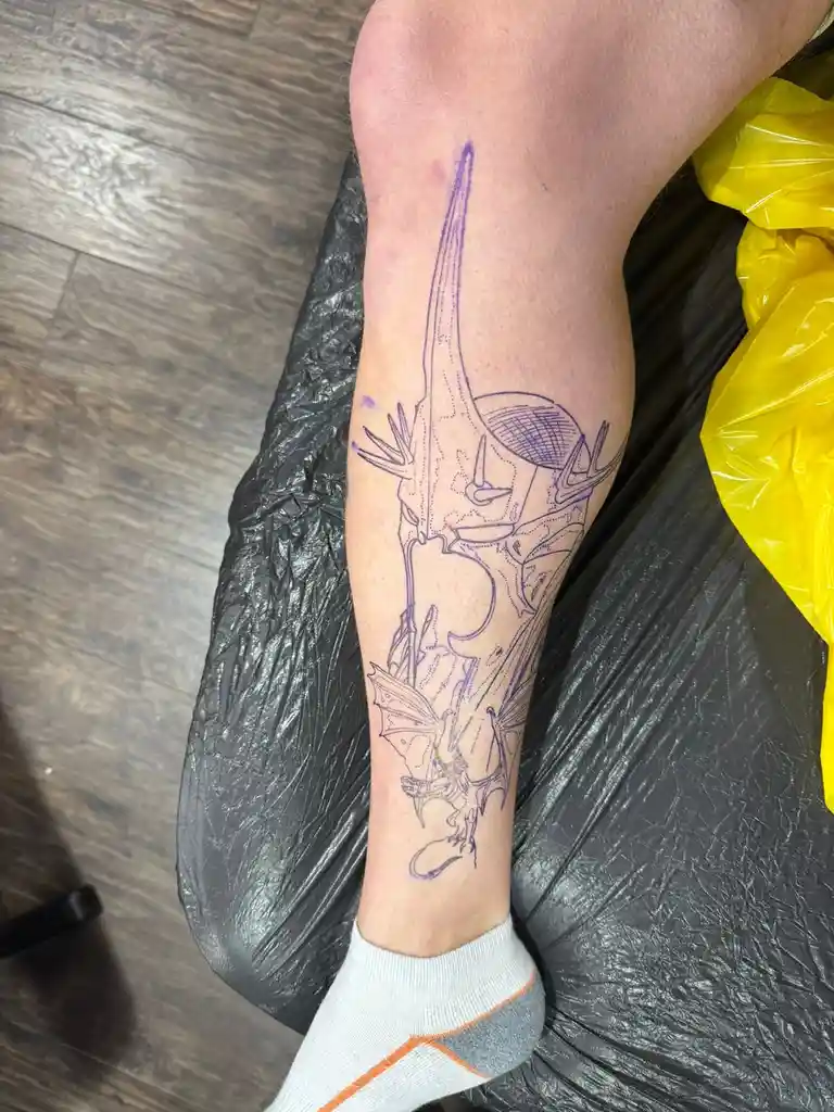 A person's lower leg and ankle, covered in a purple outline tattoo design featuring a dragon and a deer skull, rests on a…