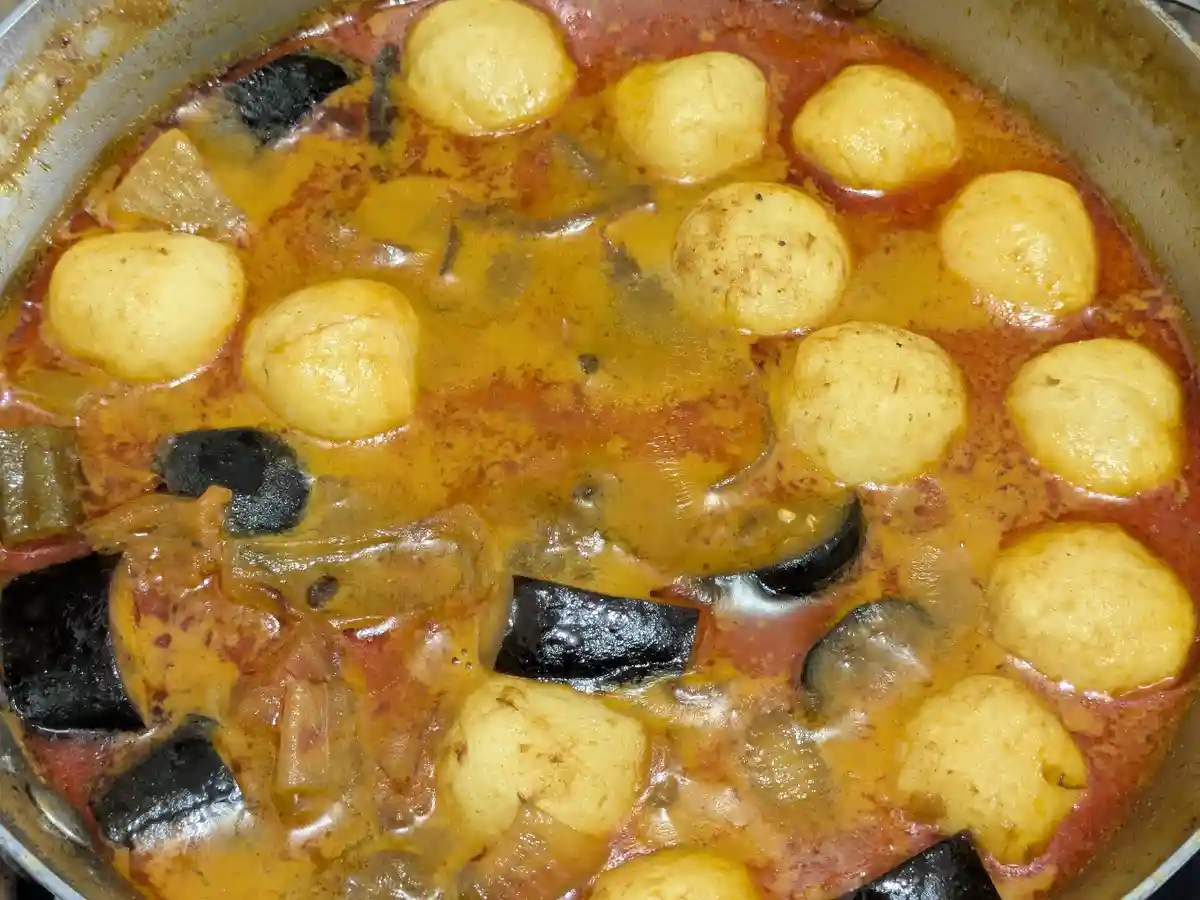 A close-up photo of a simmering curry in a metal pot, featuring numerous pale yellow, round frozen dumplings floating in a…