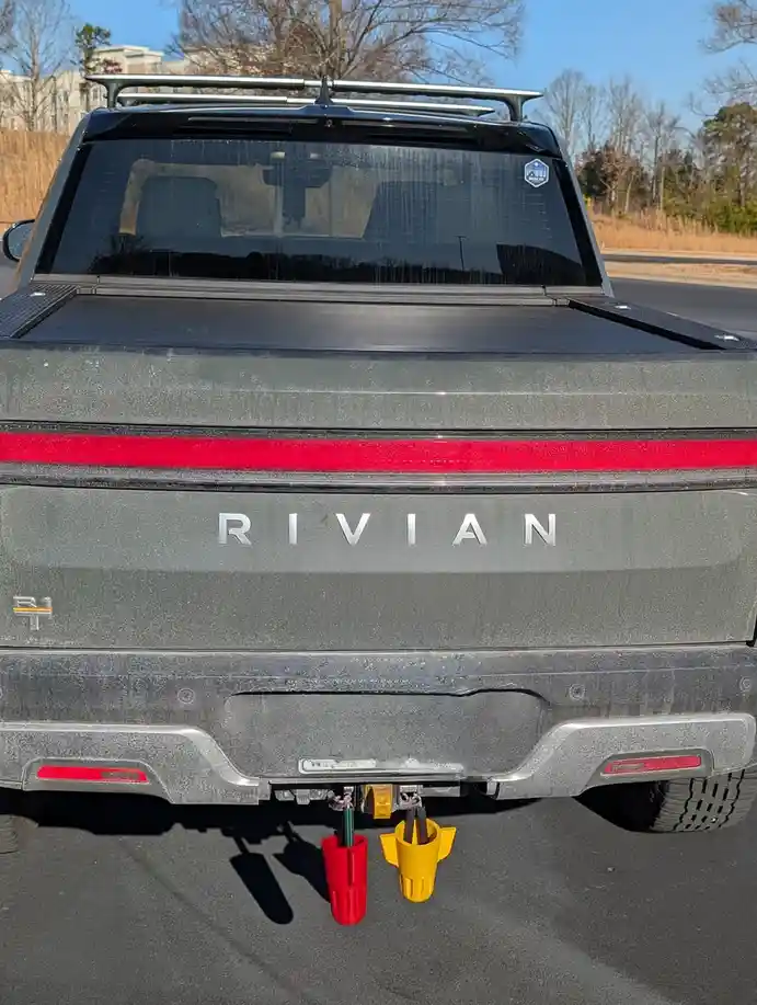 The rear view of a gray Rivian R1T electric pickup truck shows its 'RIVIAN' badge and red taillight bar. Two colorful…