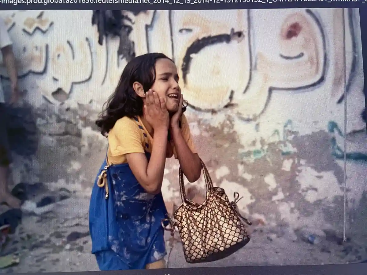 A young girl with dark hair, wearing a yellow shirt and blue overalls, stands in front of a graffiti-covered wall. She has…