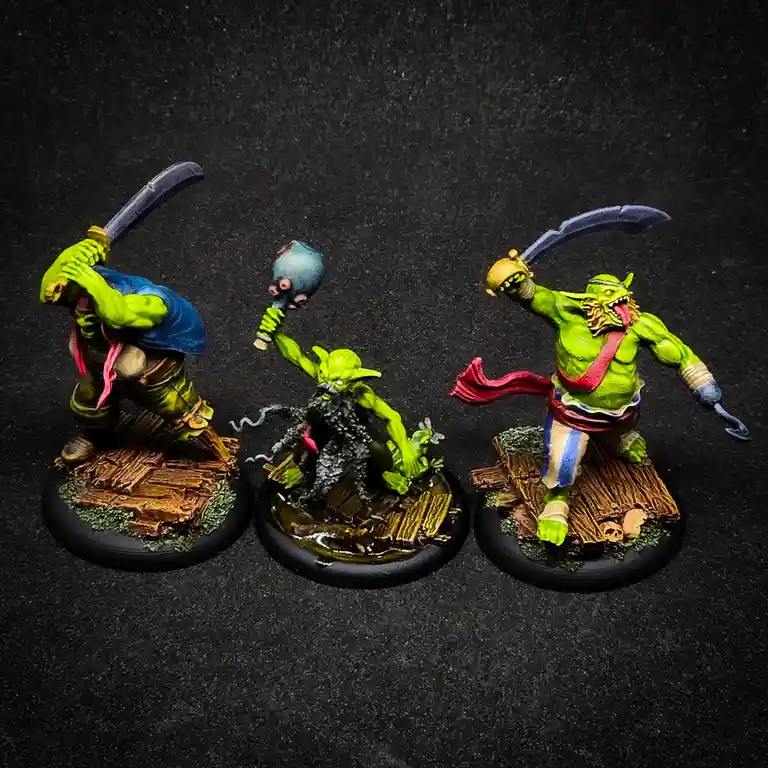 Three hand-painted green orc miniatures stand on wooden bases against a dark background. The left orc holds a curved blade…