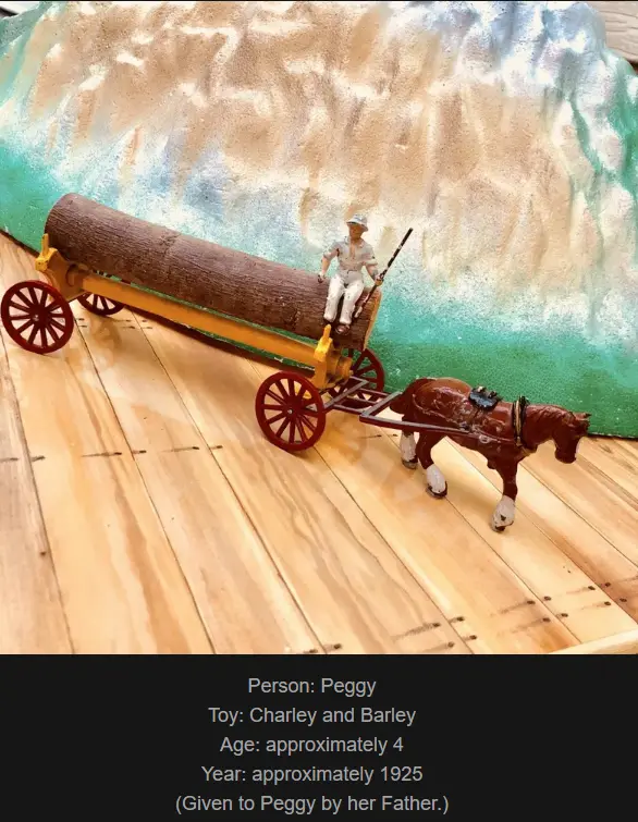 A vintage toy scene shows a small figurine of a child named Peggy sitting atop a toy log cart pulled by a toy horse, set…