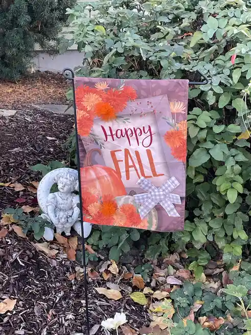 A garden flag with a 'Happy FALL' message in red and orange script, surrounded by orange flowers and pumpkins with a…