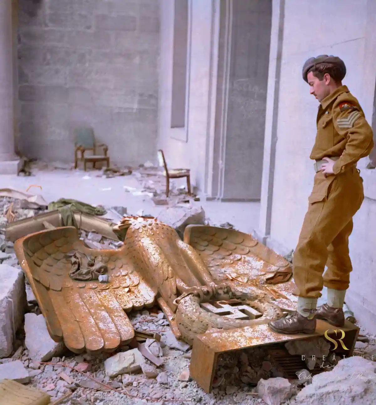 A soldier in a brown uniform stands beside a large, broken golden eagle sculpture with a swastika on its chest, amidst…