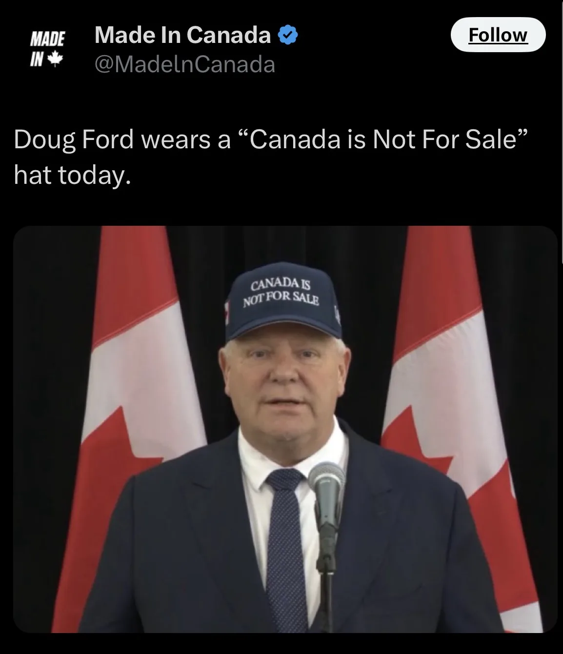 A man in a dark suit and tie stands at a podium with a microphone, wearing a blue baseball cap that reads "CANADA IS NOT FOR…