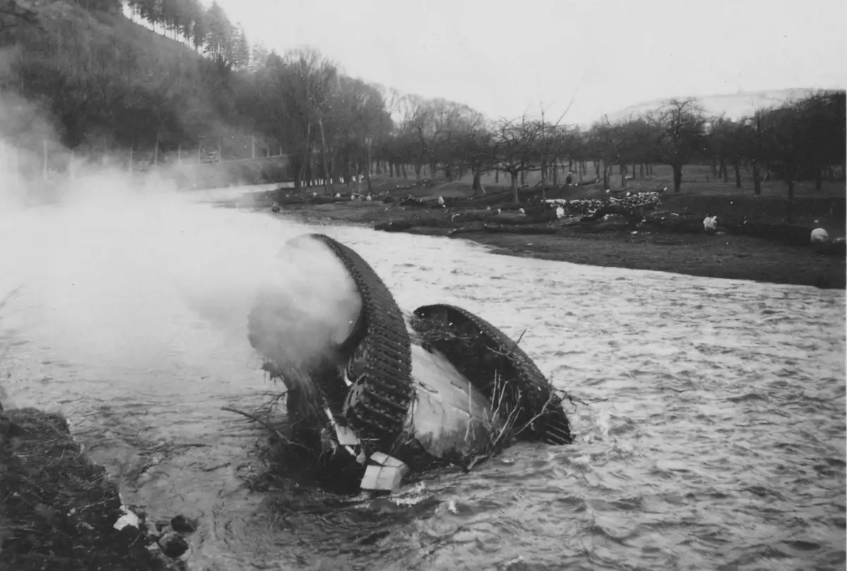 A black-and-white photo shows a tank partially submerged and tilted in a river, with smoke billowing from its side. The…
