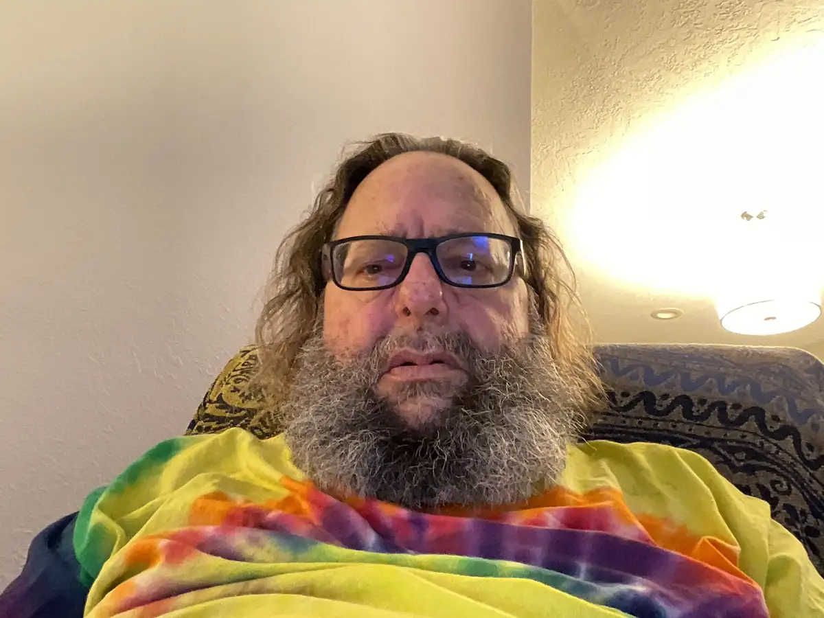 A man with a long gray beard and black-rimmed glasses sits in a chair, wrapped in a colorful rainbow tie-dye blanket. He has…