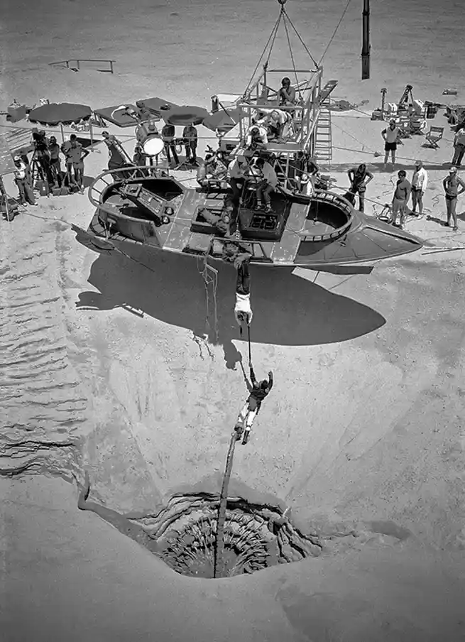 A black-and-white photo shows a large, futuristic-looking spaceship hovering over a massive hole in a sandy beach, with…