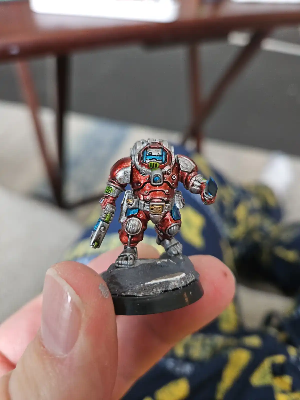 A hand holds a painted miniature figure of a heavily armored, red-and-silver robotic-looking warrior with glowing blue eyes…