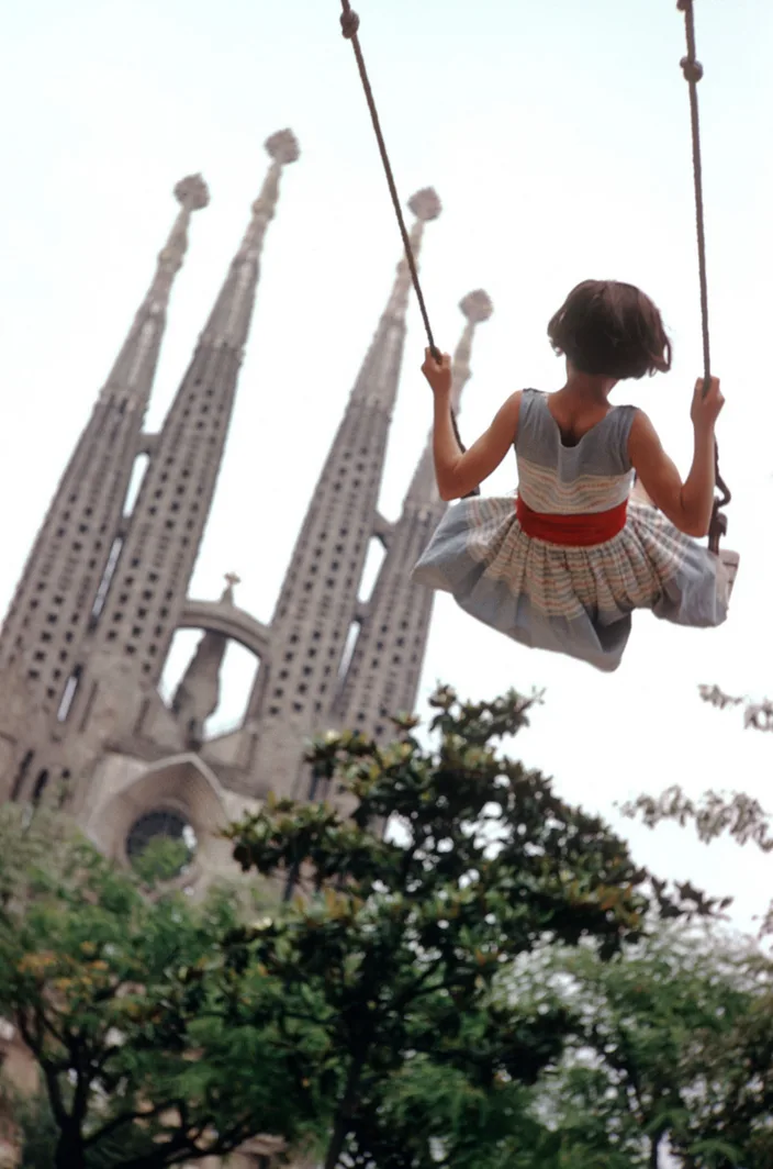 a young girl in a striped dress with a red belt is seen from behind swinging on a rope swing, with the iconic spires of…