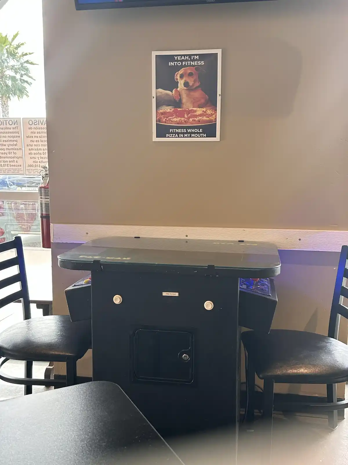 A photo of a Pac-Man arcade machine in a restaurant with two chairs, above which hangs a framed poster of a dog with a whole…
