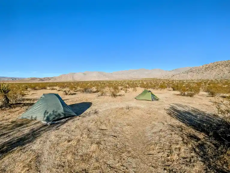 Two green tents are pitched in a vast, arid desert landscape under a clear blue sky, surrounded by sparse desert vegetation…