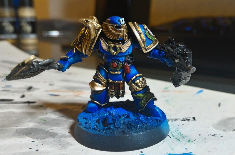 A painted miniature of a blue-armored warrior with gold accents, holding weapons, stands on a blue-painted base. The image…