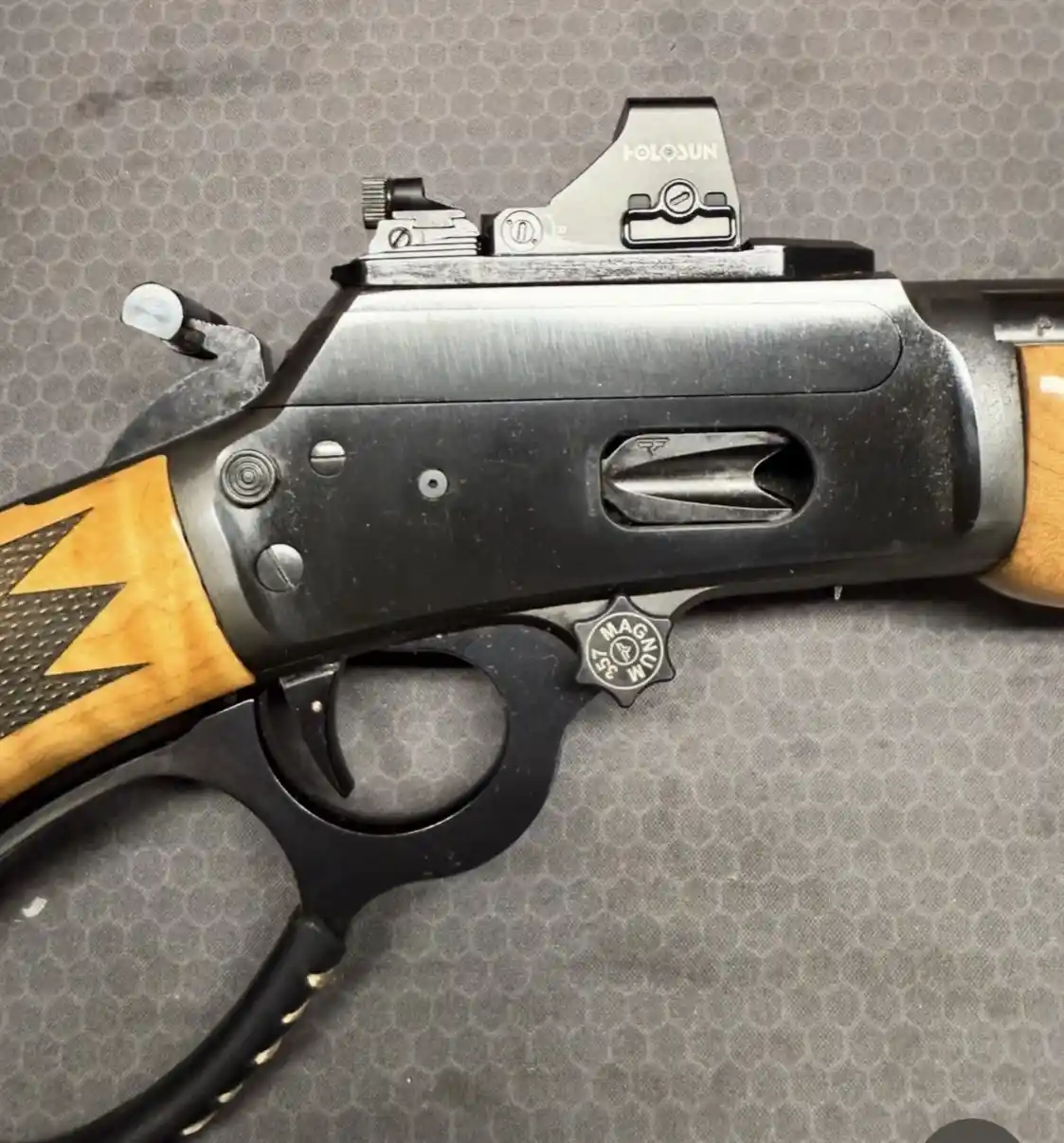 A close-up photo of a black lever-action rifle with wooden grips and a Holosun red dot sight mounted on top. The rifle has a…