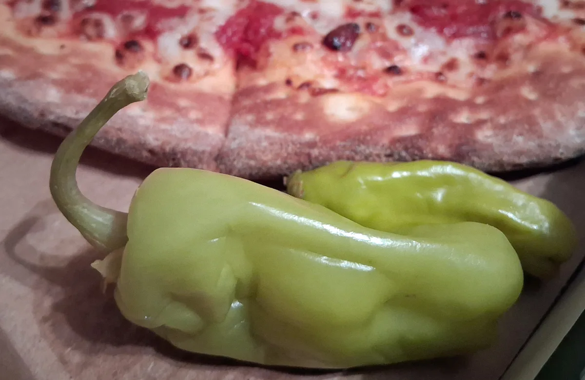 A close-up photo shows two green, glossy, wrinkled peppers in the foreground, with a blurry pizza in the background. The…