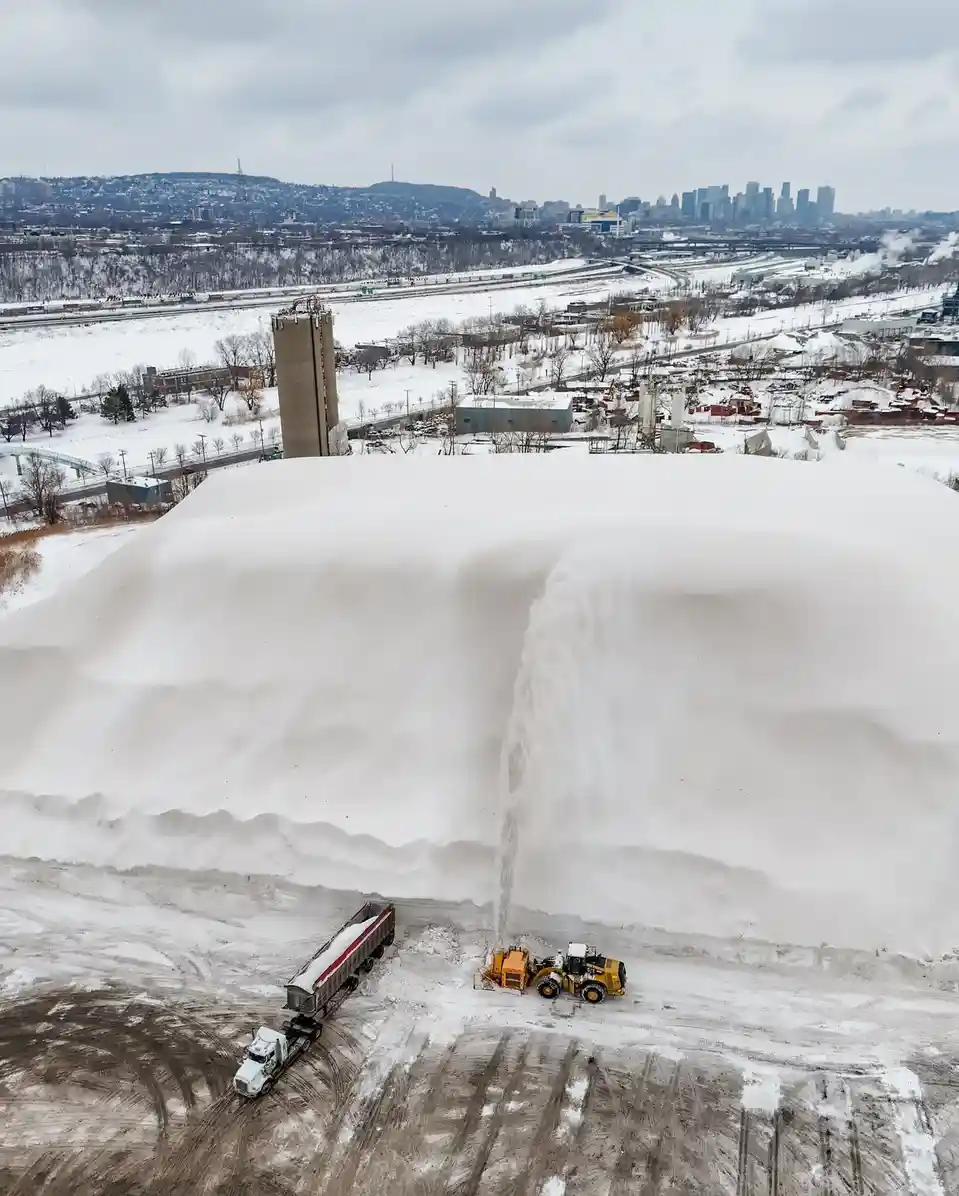 An aerial photo shows a massive snow pile being actively shoveled by a yellow front loader, sending a plume of snow into the…