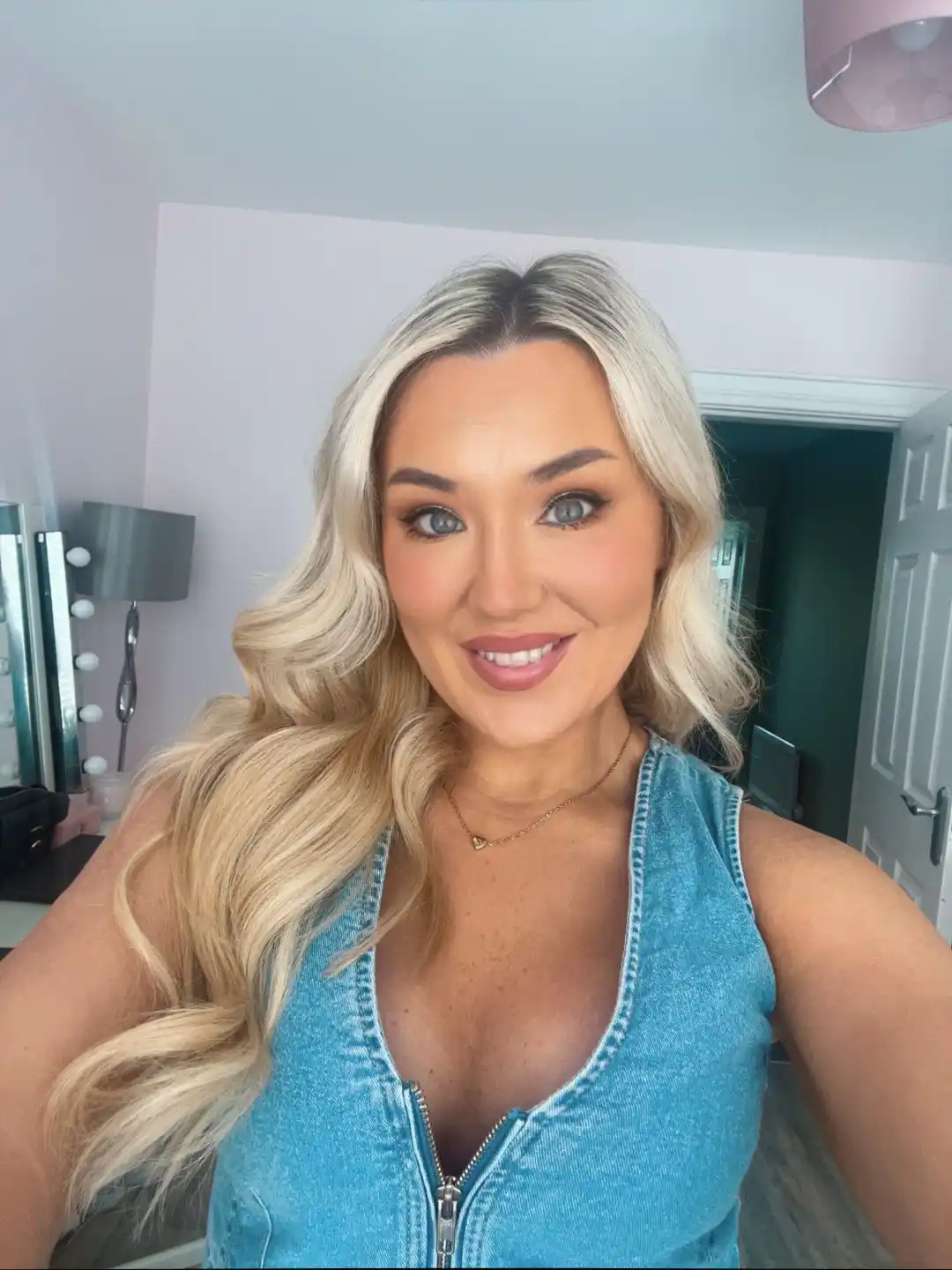 A woman with long blonde hair and makeup smiles at the camera while wearing a blue denim zip-up top. Behind her is a room…