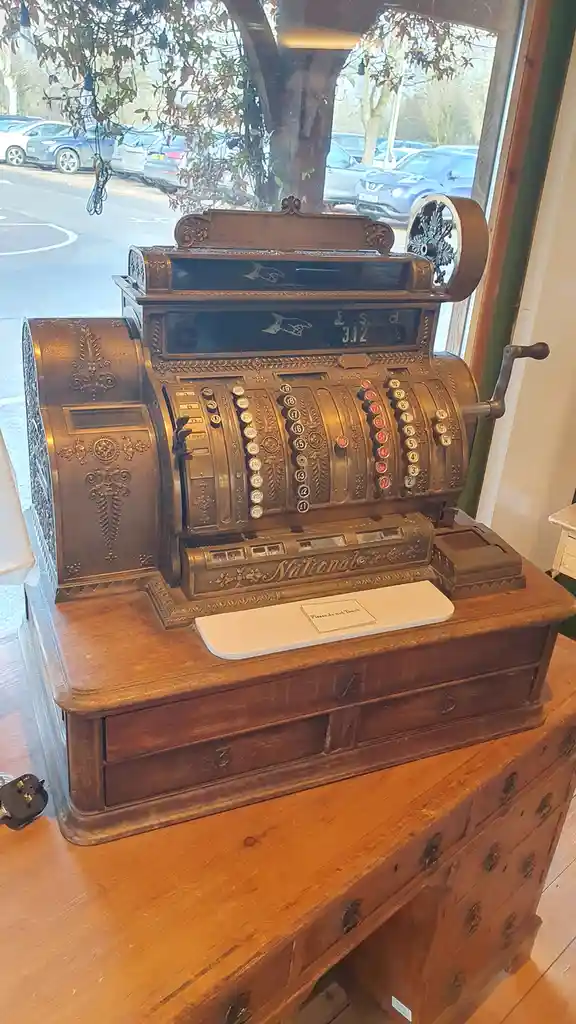 An ornate, antique National brand mechanical cash register sits on a wooden cabinet inside a shop, with a small sign on its…