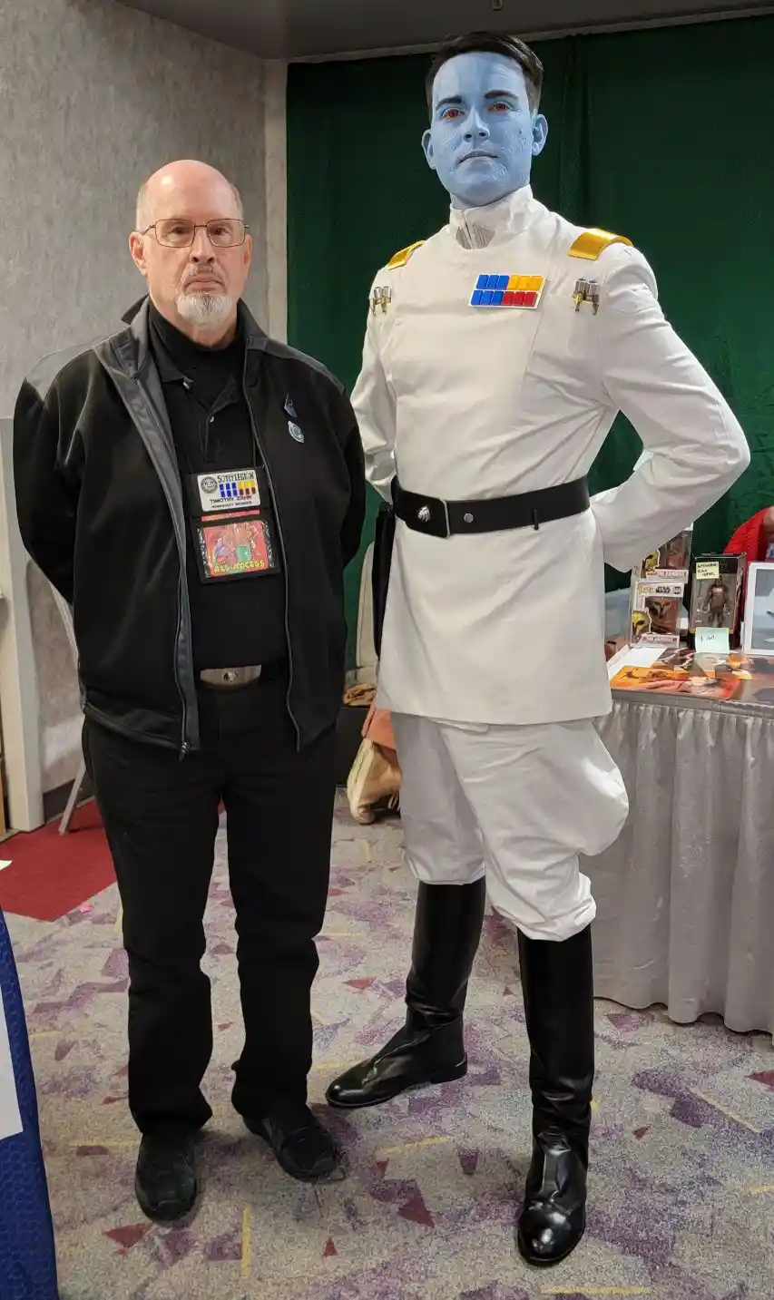 A man in a black jacket and glasses stands next to a tall person in a full-body blue-skinned Star Wars Imperial officer…