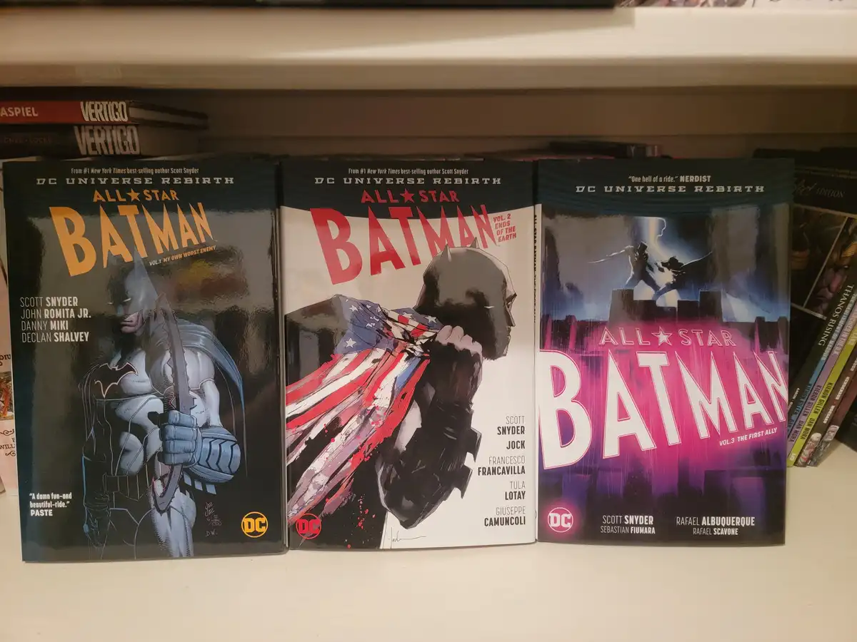 Three 'All-Star Batman' comic book volumes from the DC Universe Rebirth series are displayed side-by-side on a shelf. The…