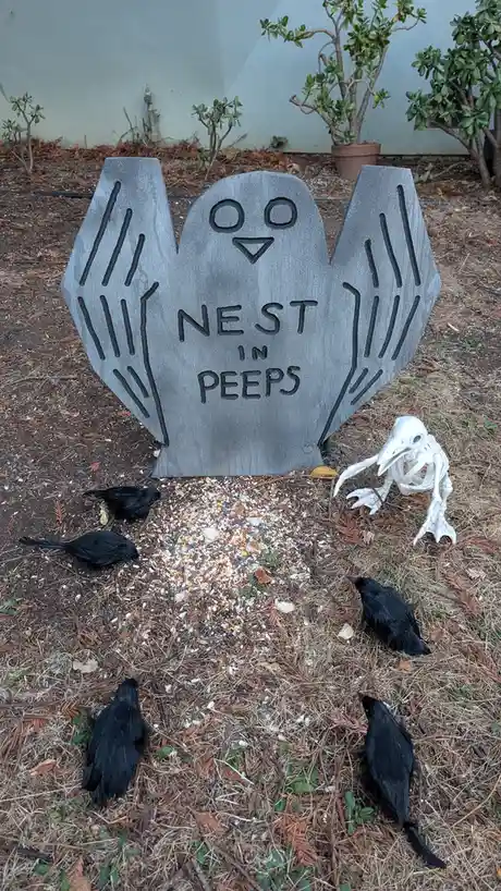 A gray, owl-shaped Halloween tombstone with carved eyes and wings stands in a yard, bearing the text 'NEST IN PEEPS'. Around…