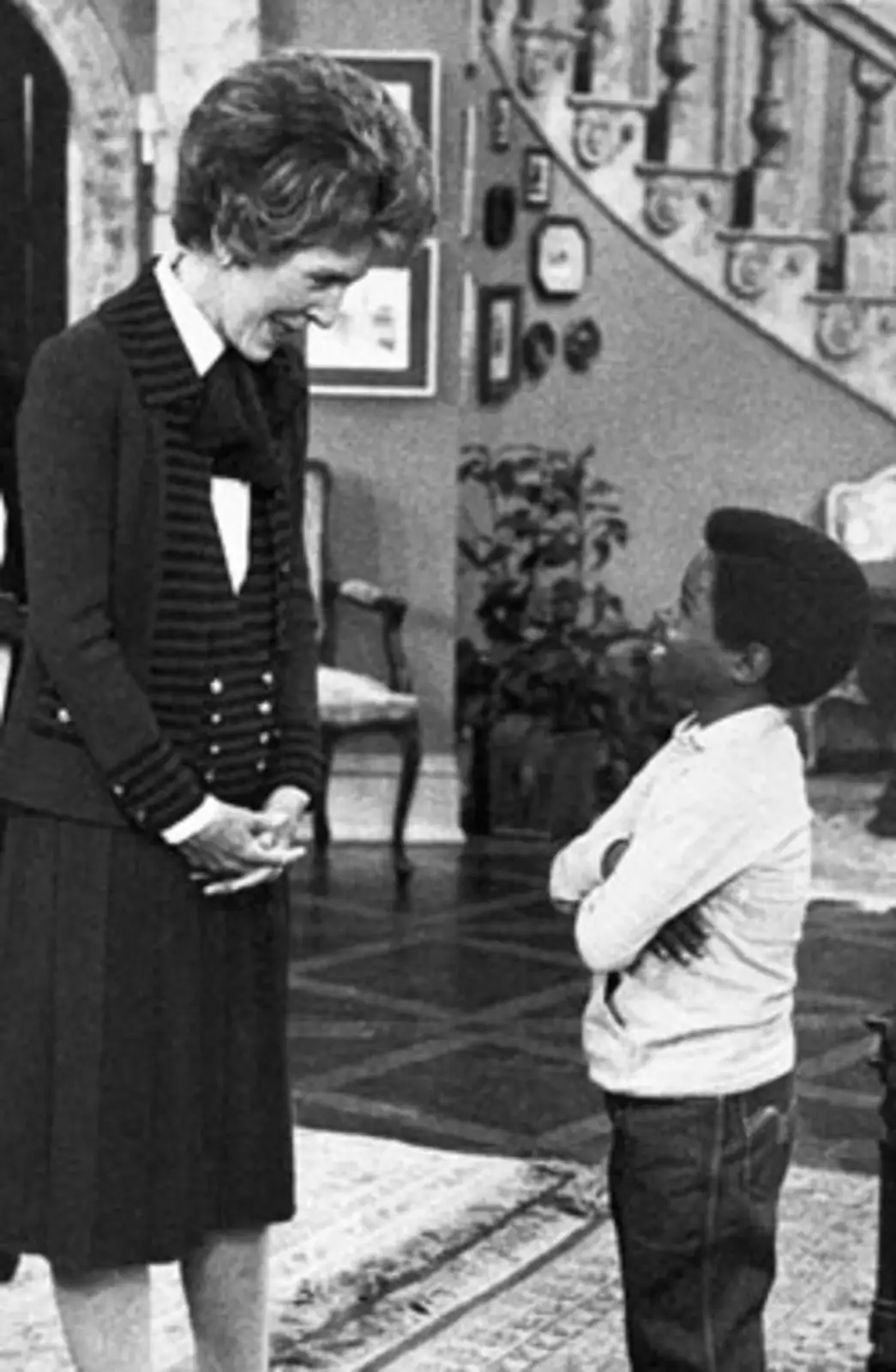 A black-and-white photo shows a woman in a dark suit smiling down at a young boy who stands with arms crossed, looking up at…